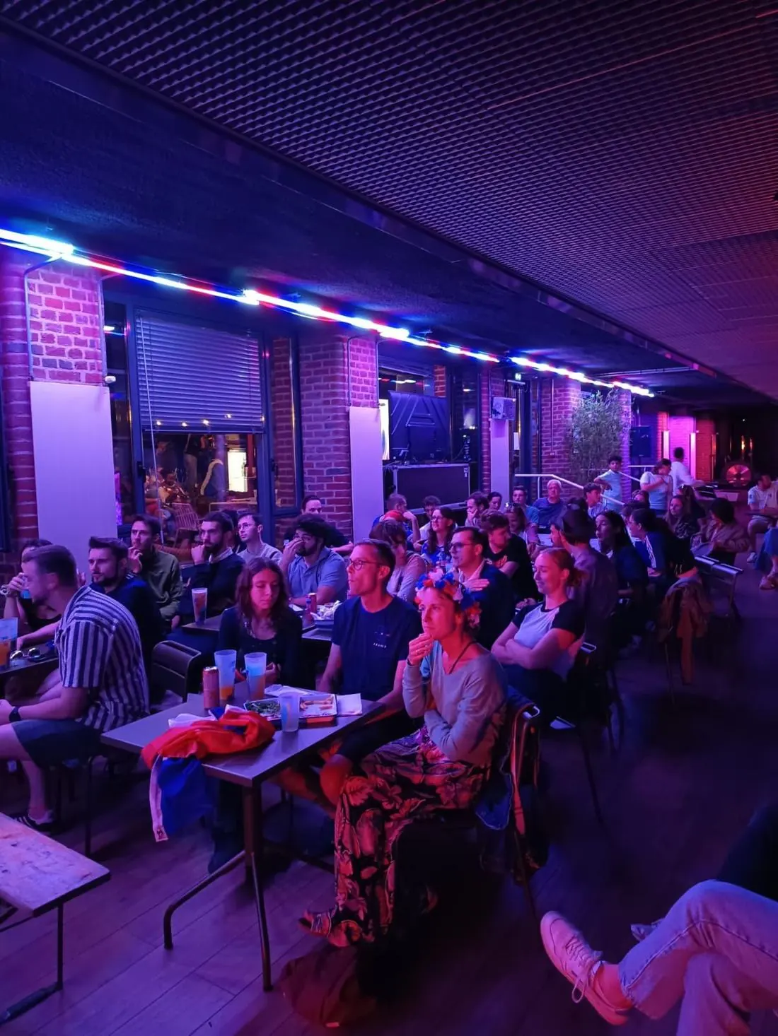 Public réuni en intérieur sous un éclairage bleu et violet pour suivre un match de football sur écran