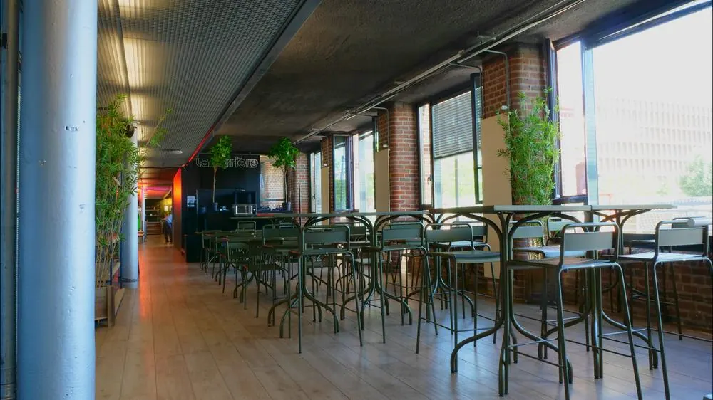 Rangée de tables hautes et de tabourets verts près des baies vitrées, dans la salle intérieure du Gong